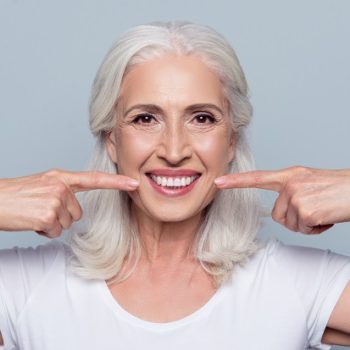 Concept of having strong healthy straight white teeth at old age. Close up portrait of happy with beaming smile female pensioner pointing on her perfect clear white teeth, isolated on gray background